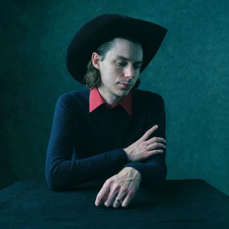 Man wearing a cowboy hat, dark sweater, and red shirt posing against a textured green background.