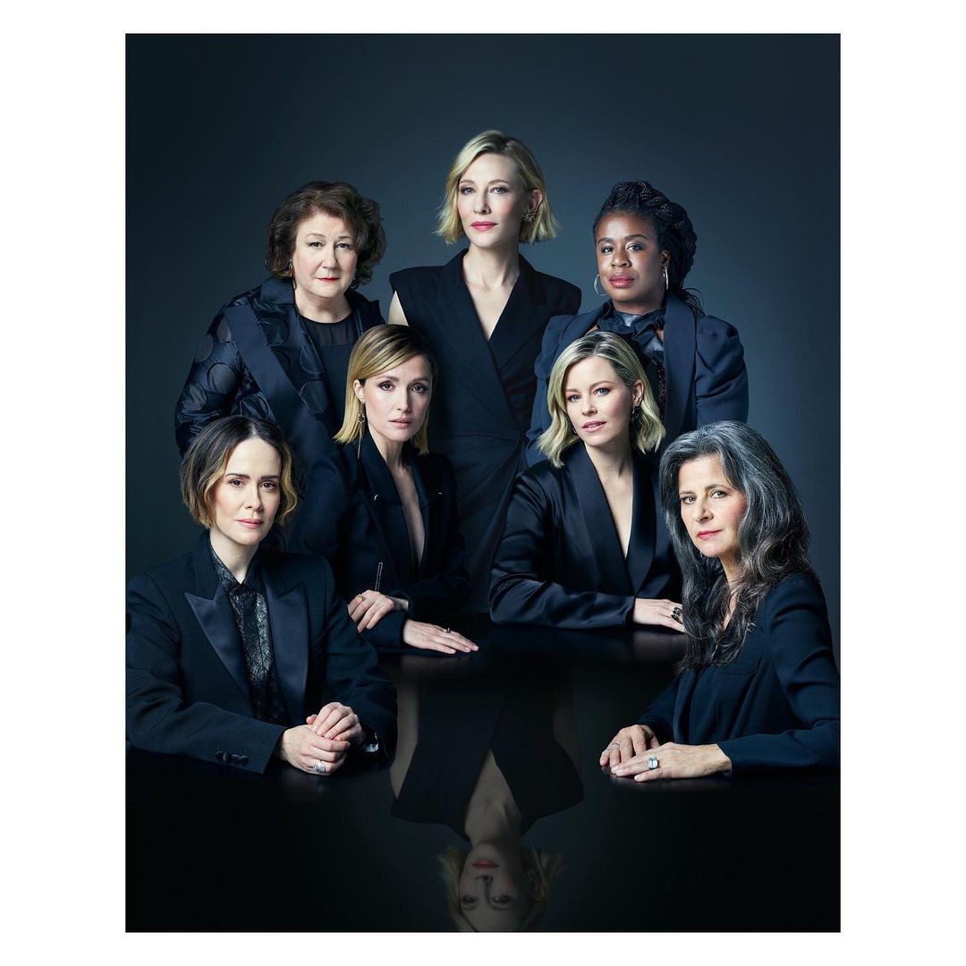 Group portrait of seven adult women in formal attire posing behind a reflective black table against a dark grey gradient background.