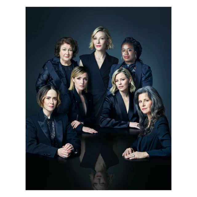 Group portrait of seven adult women in formal attire posing behind a reflective black table against a dark grey gradient background.