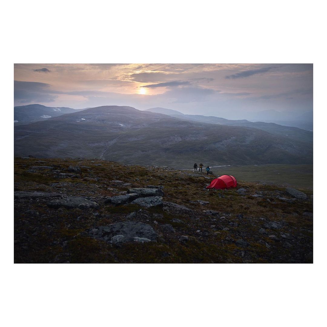 A red tent sits on a rocky plain under a cloudy sky with a group of four hikers standing off in the distance on a hillside during daytime.