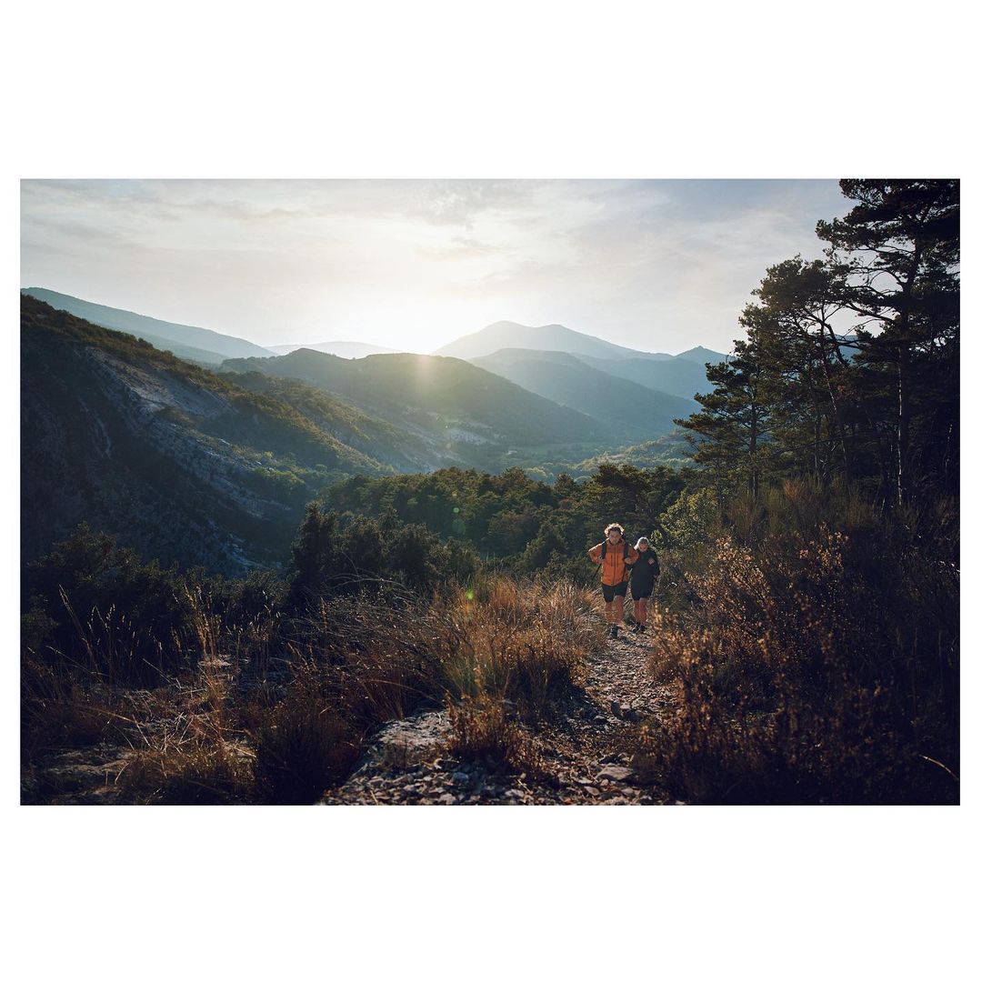Two people hike on a mountain trail amid a forest landscape under bright sunlight, with views of distant mountains and trees.