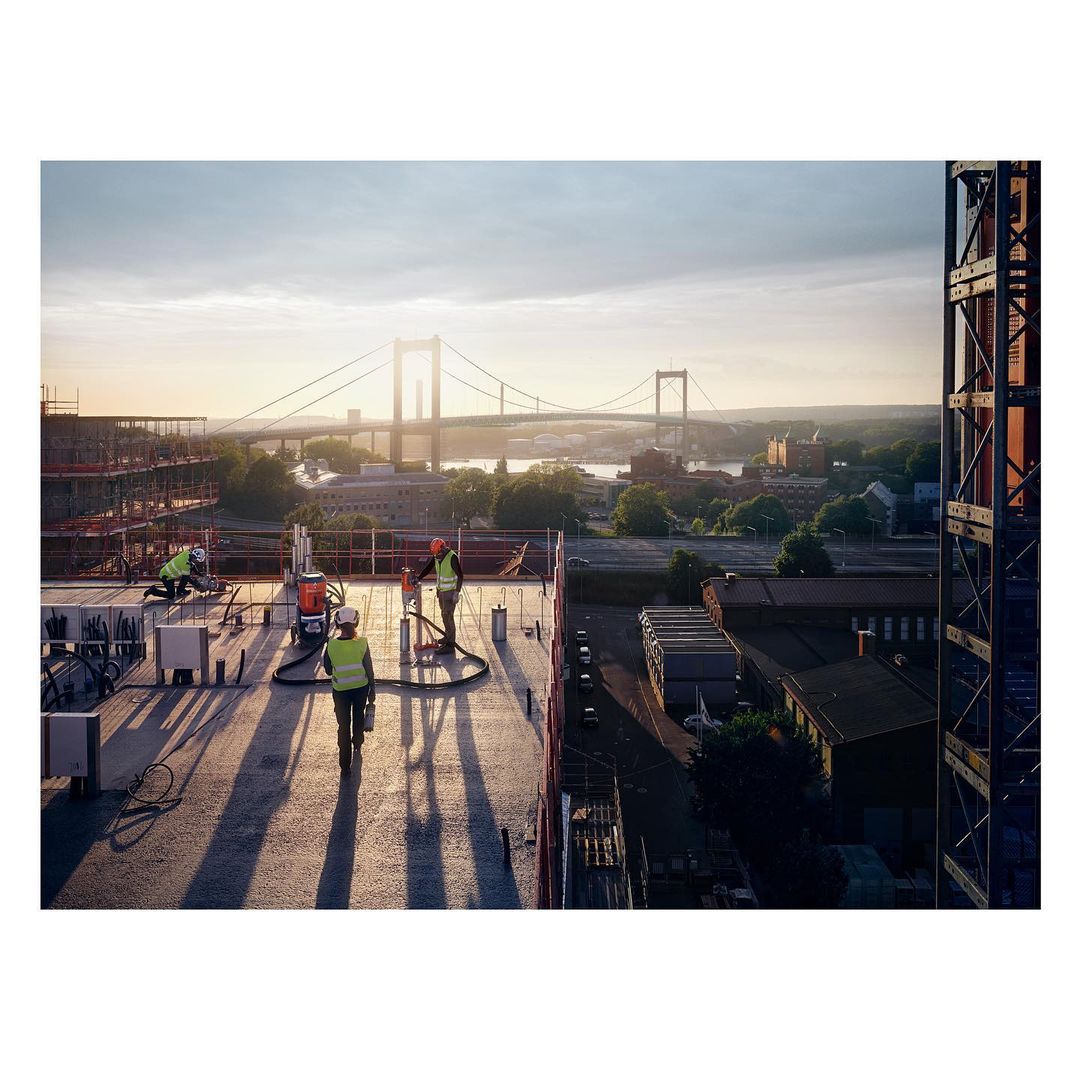 Construction workers on a concrete rooftop at sunset with a bridge and city skyline in the background. Construction crane visible.