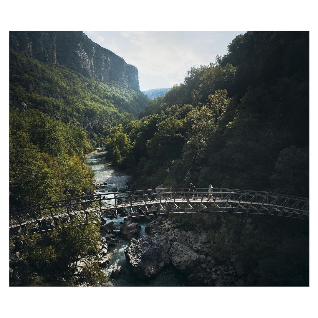 Scenic view of two people walking across a wooden bridge over a flowing river surrounded by a lush, green forest and mountains.
