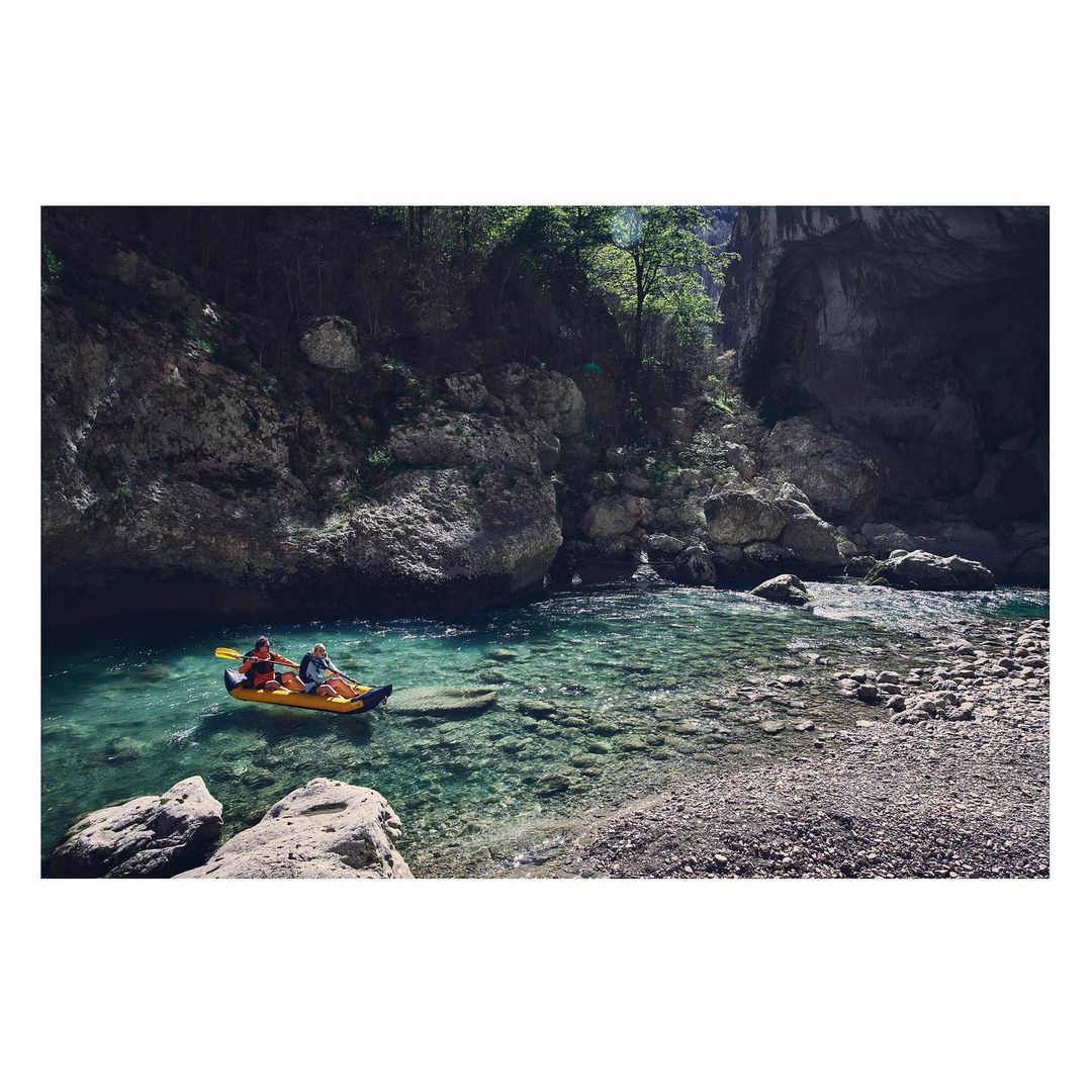 Two people paddle an inflatable yellow kayak on clear, turquoise water in a rocky river gorge with green trees and boulders.