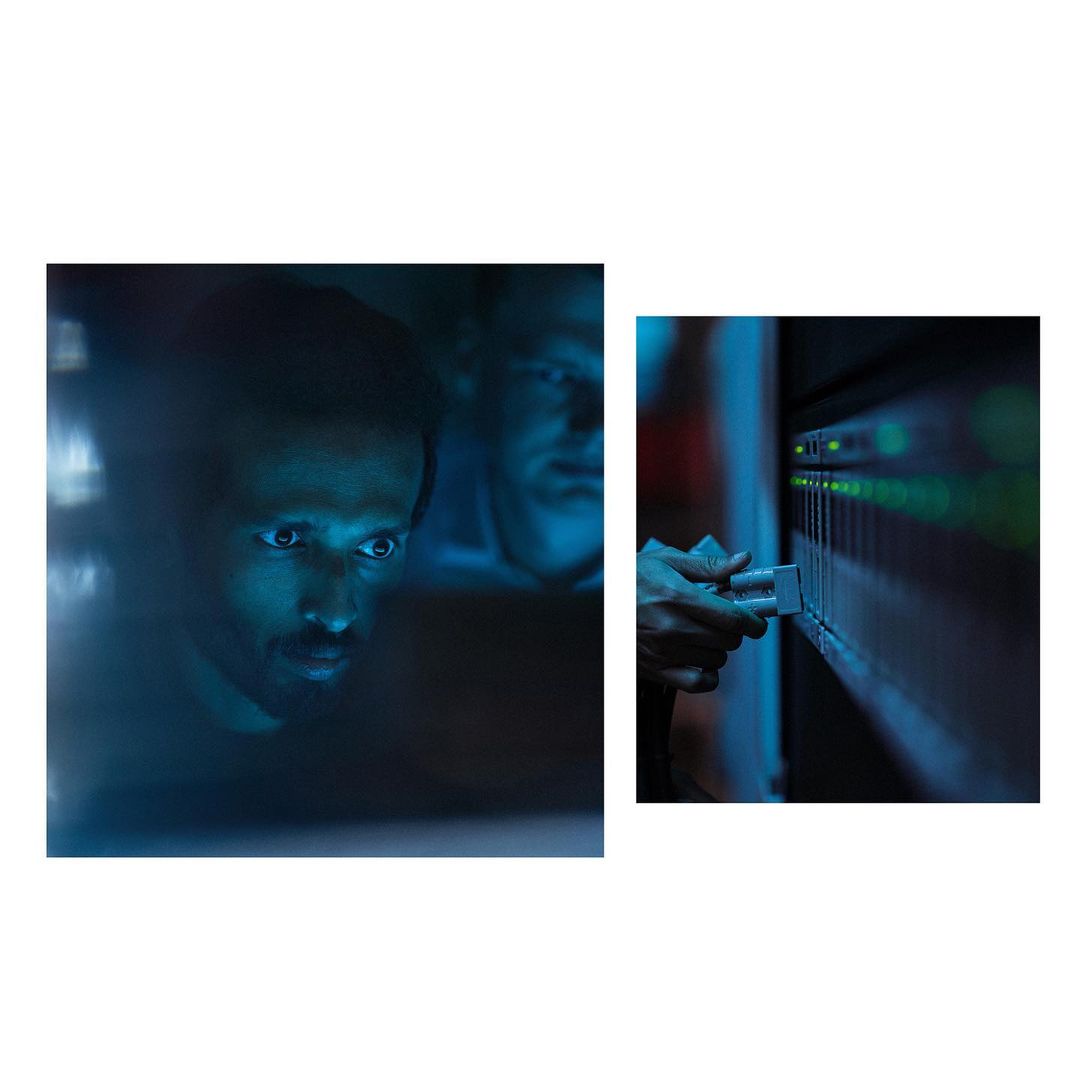 Two men in a dimly lit server room, one plugs in a server rack cable. Low-angle view with the men and equipment highlighted by blue tones.