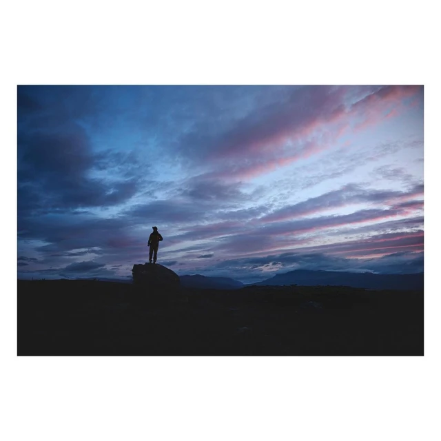 Silhouette of a person standing on a rock, observing a dramatic, colorful sunset sky over a dark, hilly landscape.
