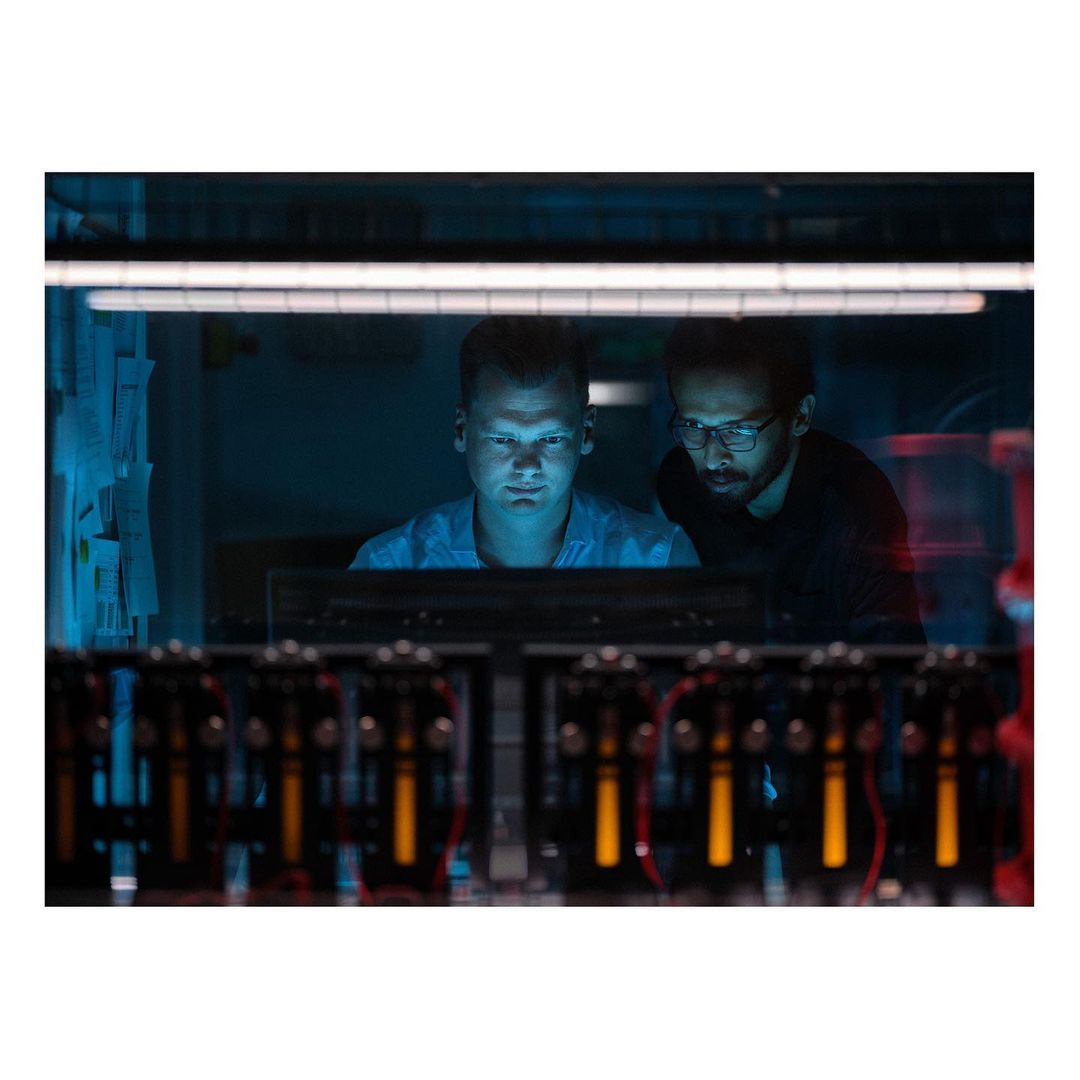 Two men are illuminated by the blue light of computer screens in a tech lab, surrounded by hardware featuring red and yellow cables.