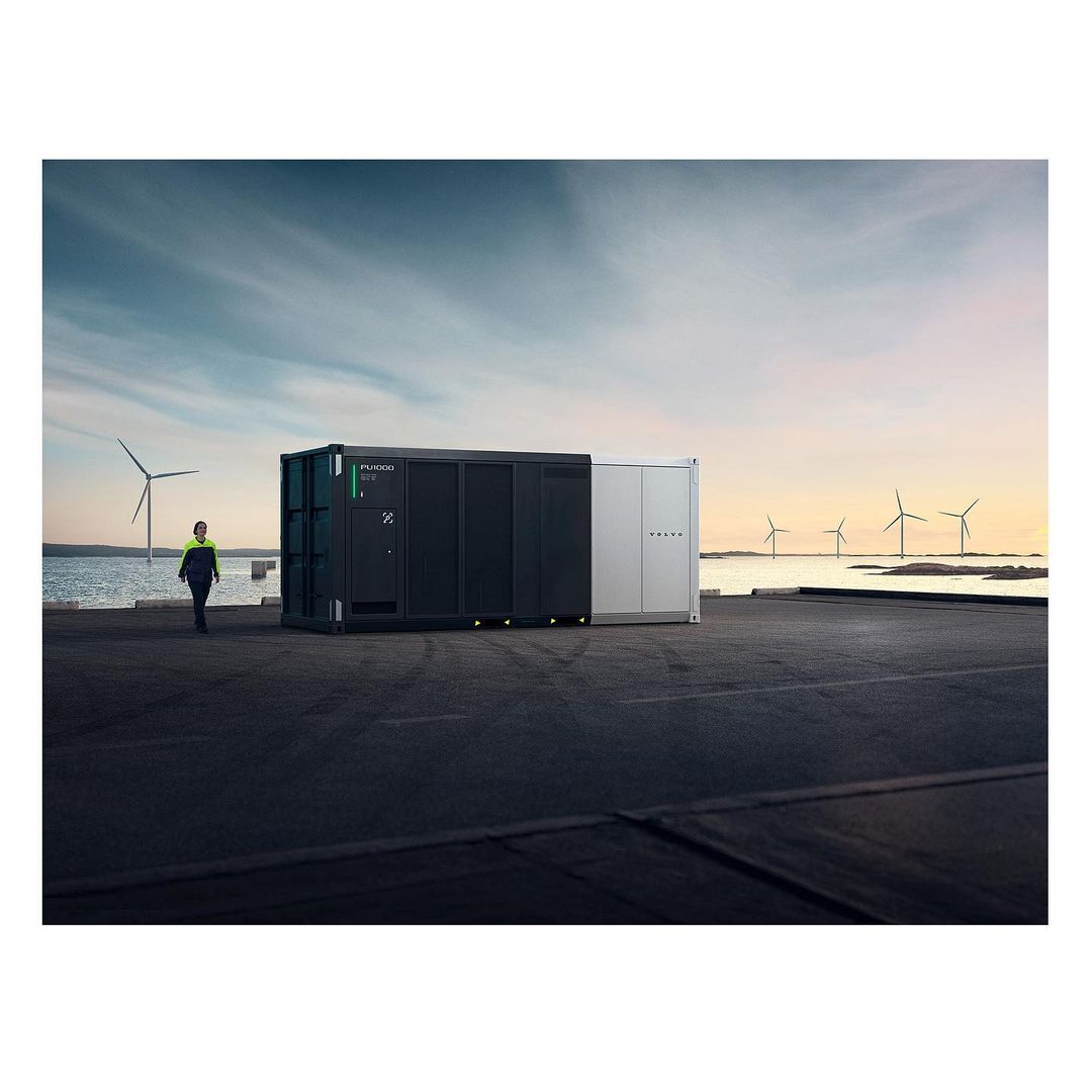 Shipping container-style energy storage system on concrete surface with wind turbines in the background, woman walking, overcast sky.
