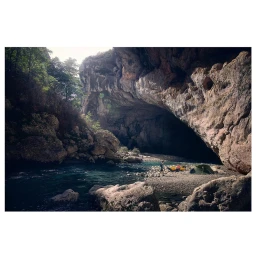 Campsite by a river underneath a massive rocky overhang with towering cliffs and trees under natural light