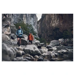 Two hikers walking along a rocky riverbed beneath towering stone cliffs, photographed by Robert Elmengård.