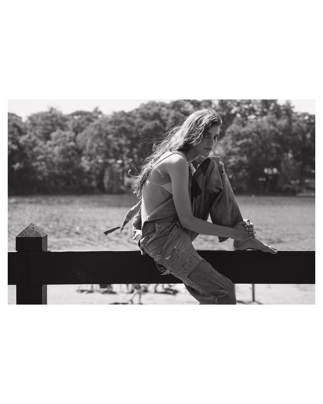 Young woman with long hair sits on a wooden fence wearing a denim overall with a lake and trees in the background in a black and white outdoor fashion shot.