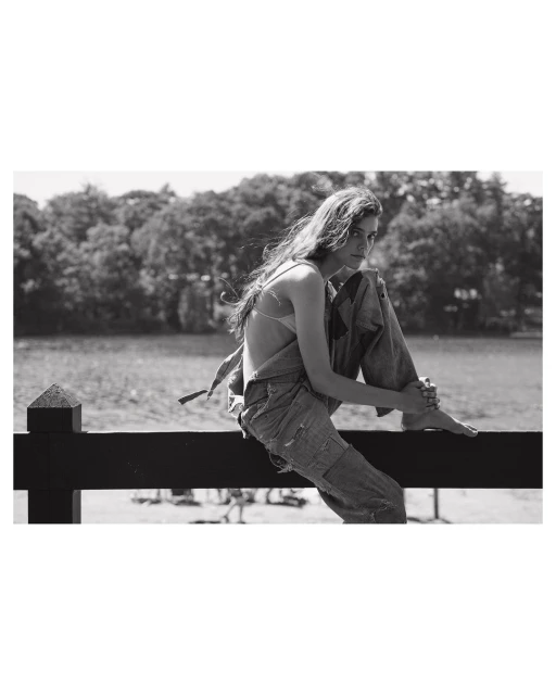 Young woman with long hair sits on a wooden fence wearing a denim overall with a lake and trees in the background in a black and white outdoor fashion shot.