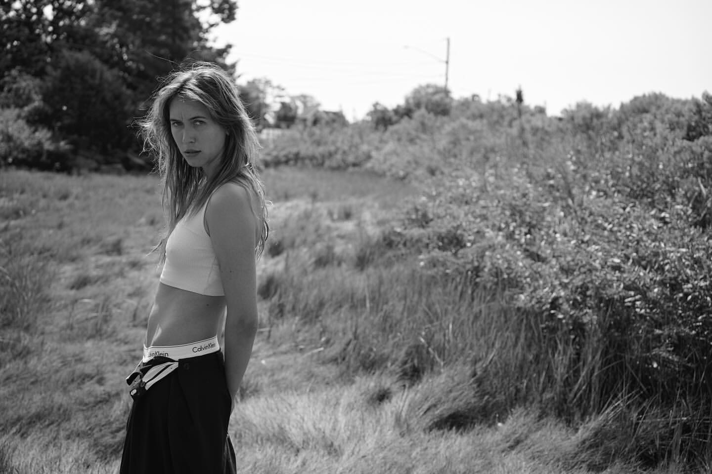 Black and white portrait of a woman in a crop top and dark trousers standing outdoors in a grassy field
