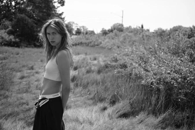 Black and white portrait of a woman in a crop top and dark trousers standing outdoors in a grassy field