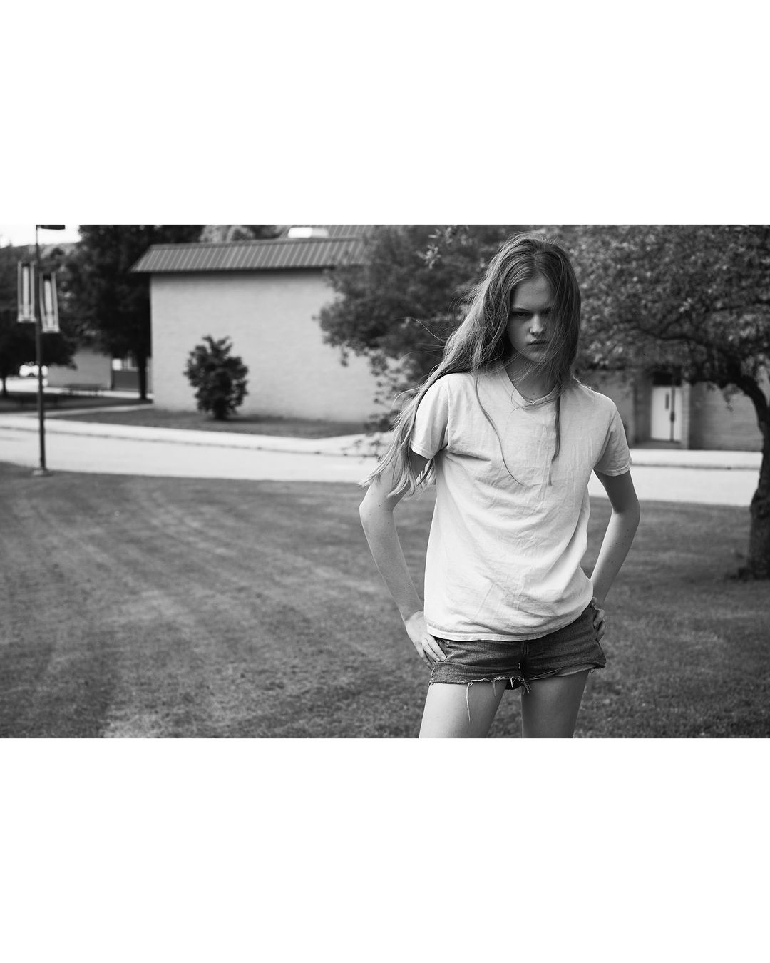 Monochrome portrait of a young adult female with long hair, standing in a grassy yard with a neutral expression, wearing casual clothing.