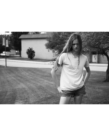 Monochrome portrait of a young adult female with long hair, standing in a grassy yard with a neutral expression, wearing casual clothing.