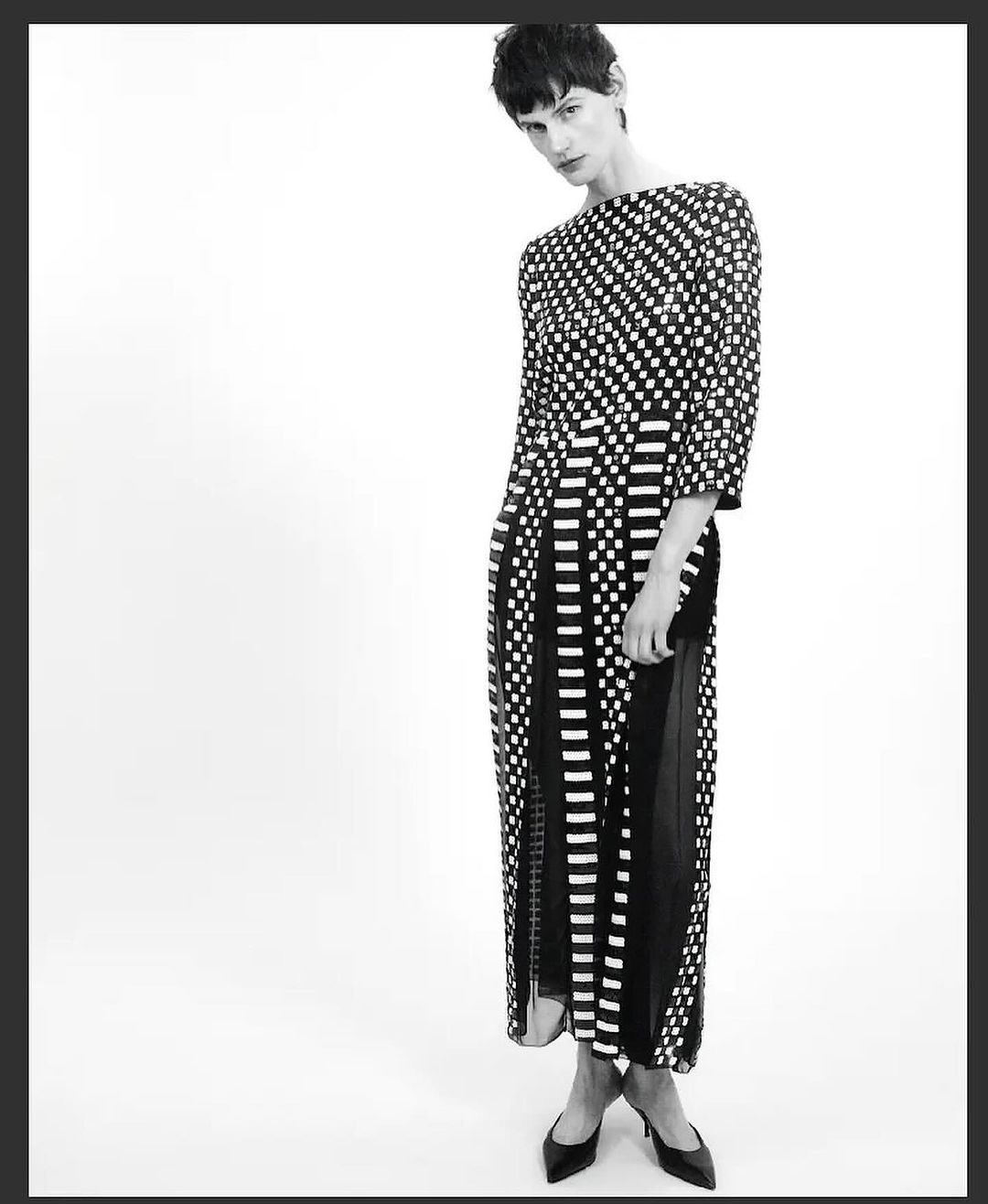 Black and white studio shot of a fashion model in a polka dot dress with slit and pumps, against a white backdrop.