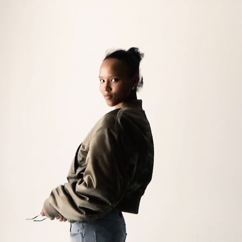 Young woman in a green bomber jacket and blue jeans looking over her shoulder against a light background in a fashion style portrait.