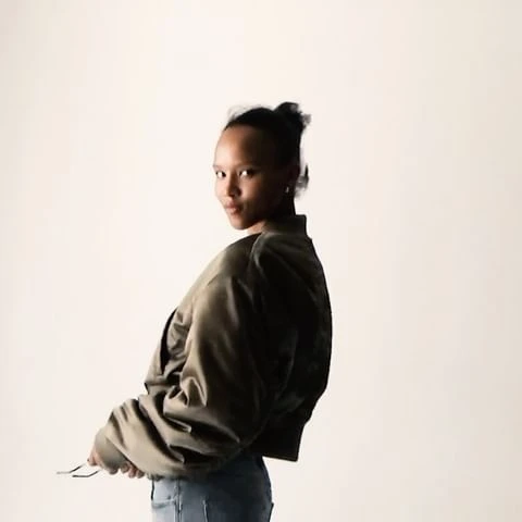 Young woman in a green bomber jacket and blue jeans looking over her shoulder against a light background in a fashion style portrait.