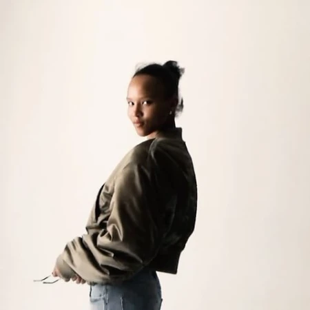 Young woman in a green bomber jacket and blue jeans looking over her shoulder against a light background in a fashion style portrait.