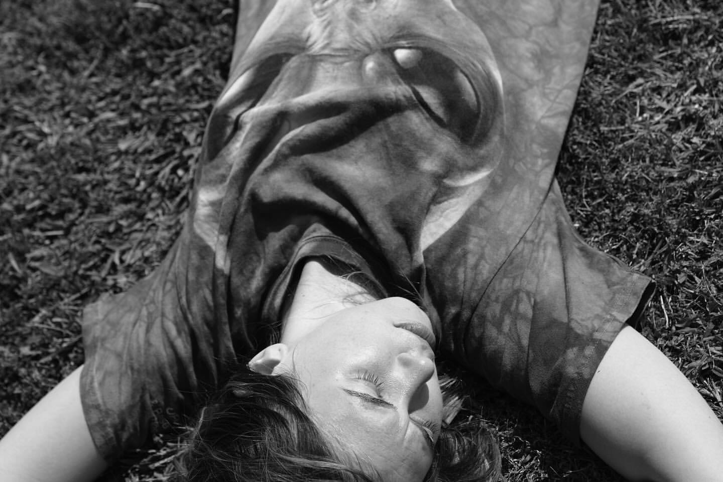 A woman lies on grass, eyes closed, wearing a graphic tee shirt in black and white in an outdoor setting with natural lighting.