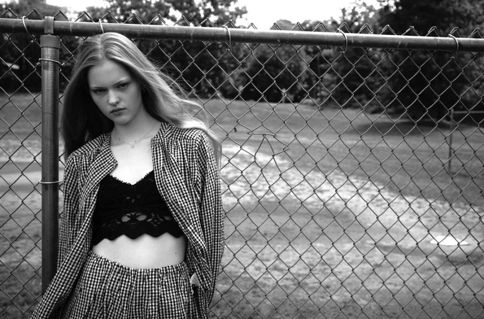 Black and white portrait of a young woman standing near a chain-link fence, wearing a bralette and a matching gingham jacket and pants.