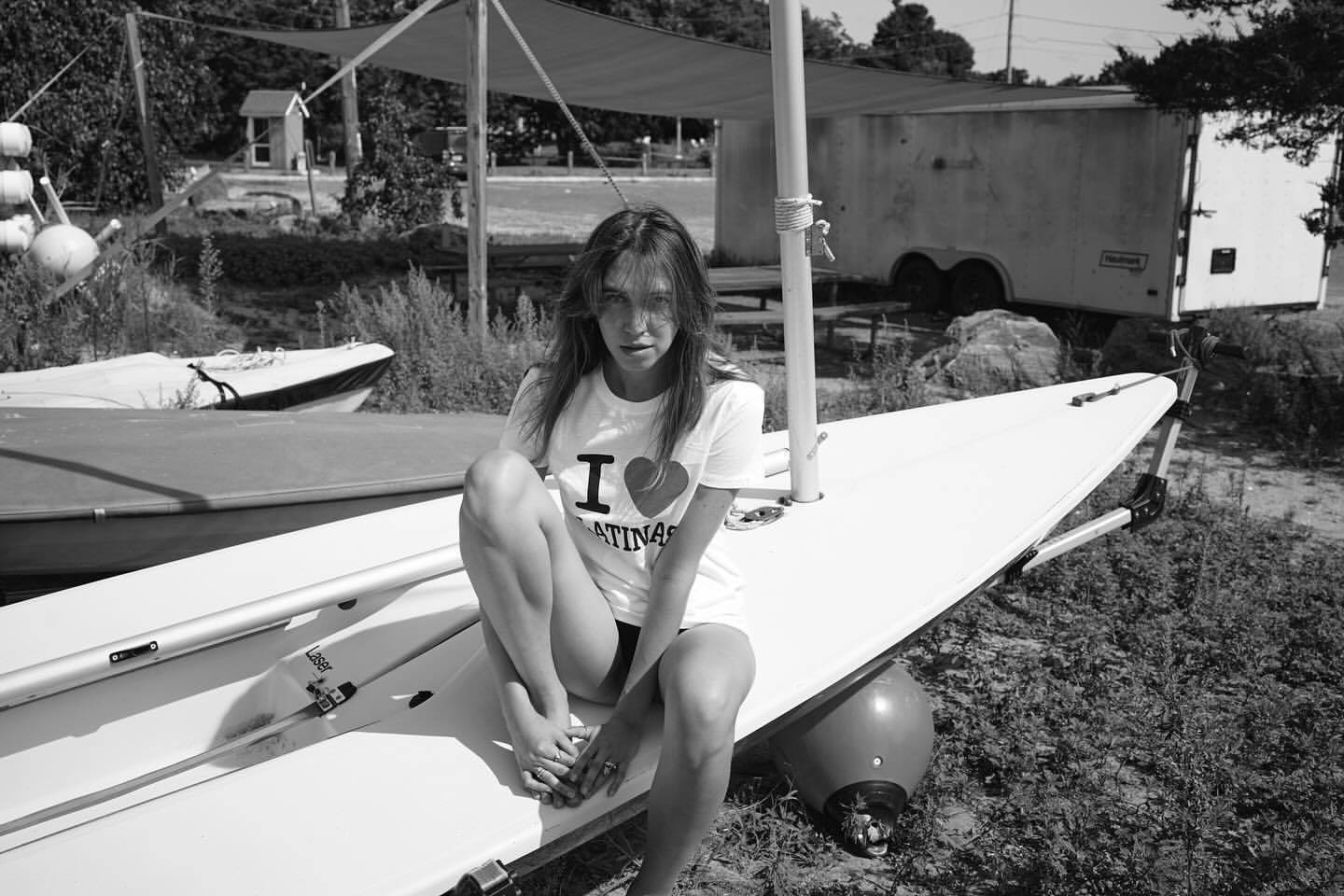 Monochrome shot of a woman with an 'I heart Latinas' tee sitting on a sailboat, with another boat and vegetation in the backdrop.