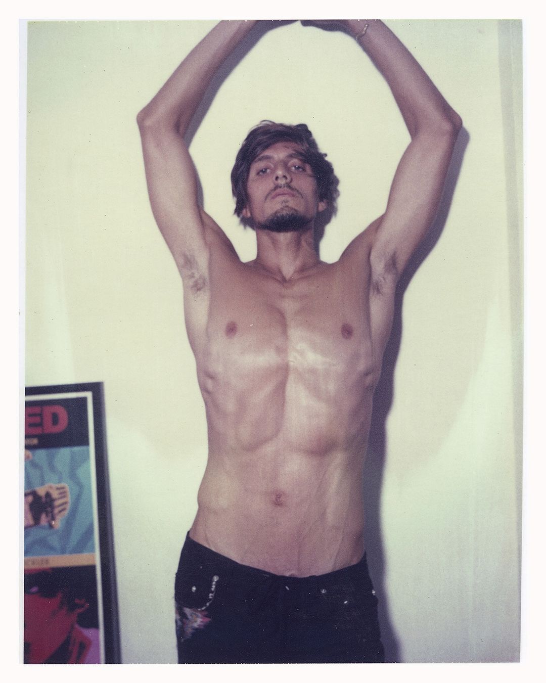 Portrait of a shirtless adult male with arms raised, wearing black pants, in front of an off-white wall with a poster.