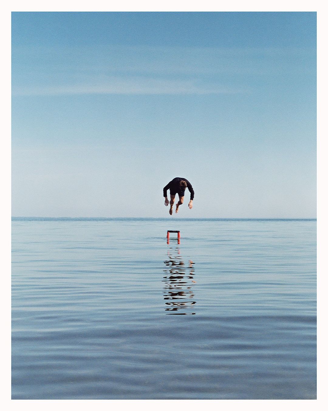 Adult male in mid-air leaping over red box in middle of wavy ocean with pale blue sky in daytime. Action shot, studio photography.