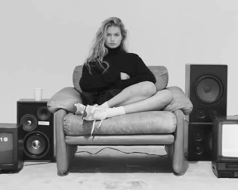 Black and white shot of a woman in a turtleneck sweater sitting in an armchair, flanked by speakers and an old television set.