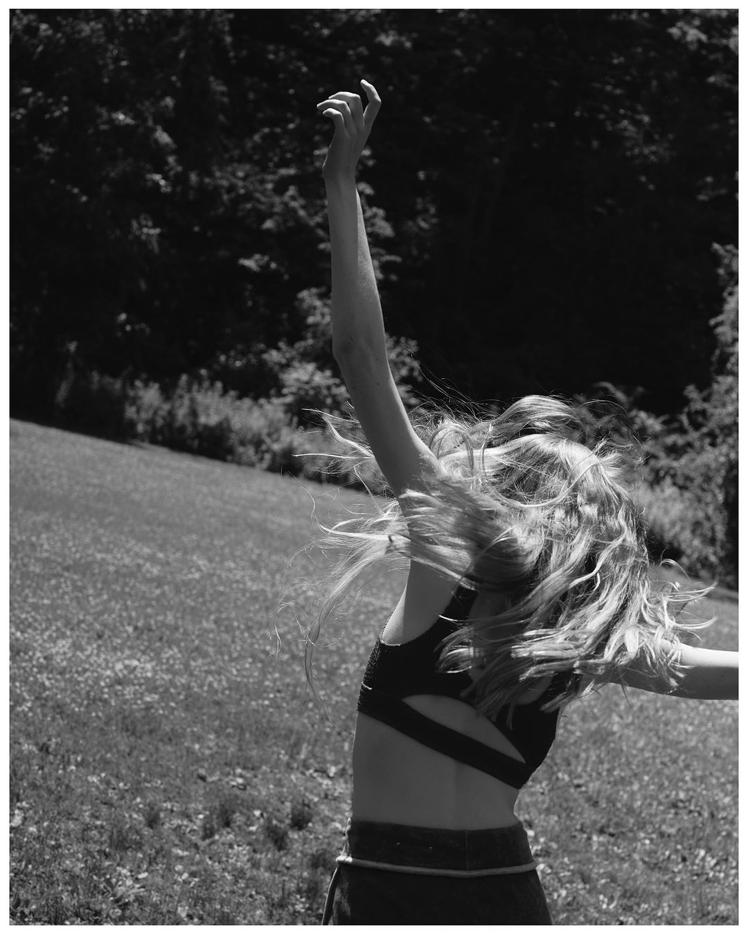 Monochrome portrait of a blonde woman with flowing hair wearing activewear in a sunlit field surrounded by trees, arms raised.