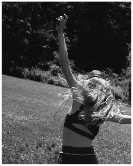 Monochrome portrait of a blonde woman with flowing hair wearing activewear in a sunlit field surrounded by trees, arms raised.