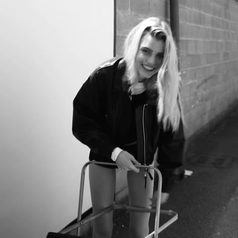 A young woman with long blonde hair smiles as she playfully uses a medical walker in a black-and-white medium shot against a wall.