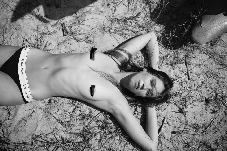 A woman lying on sandy ground in black bikini bottoms, sunlit with long hair, in a black and white outdoor fashion shot.