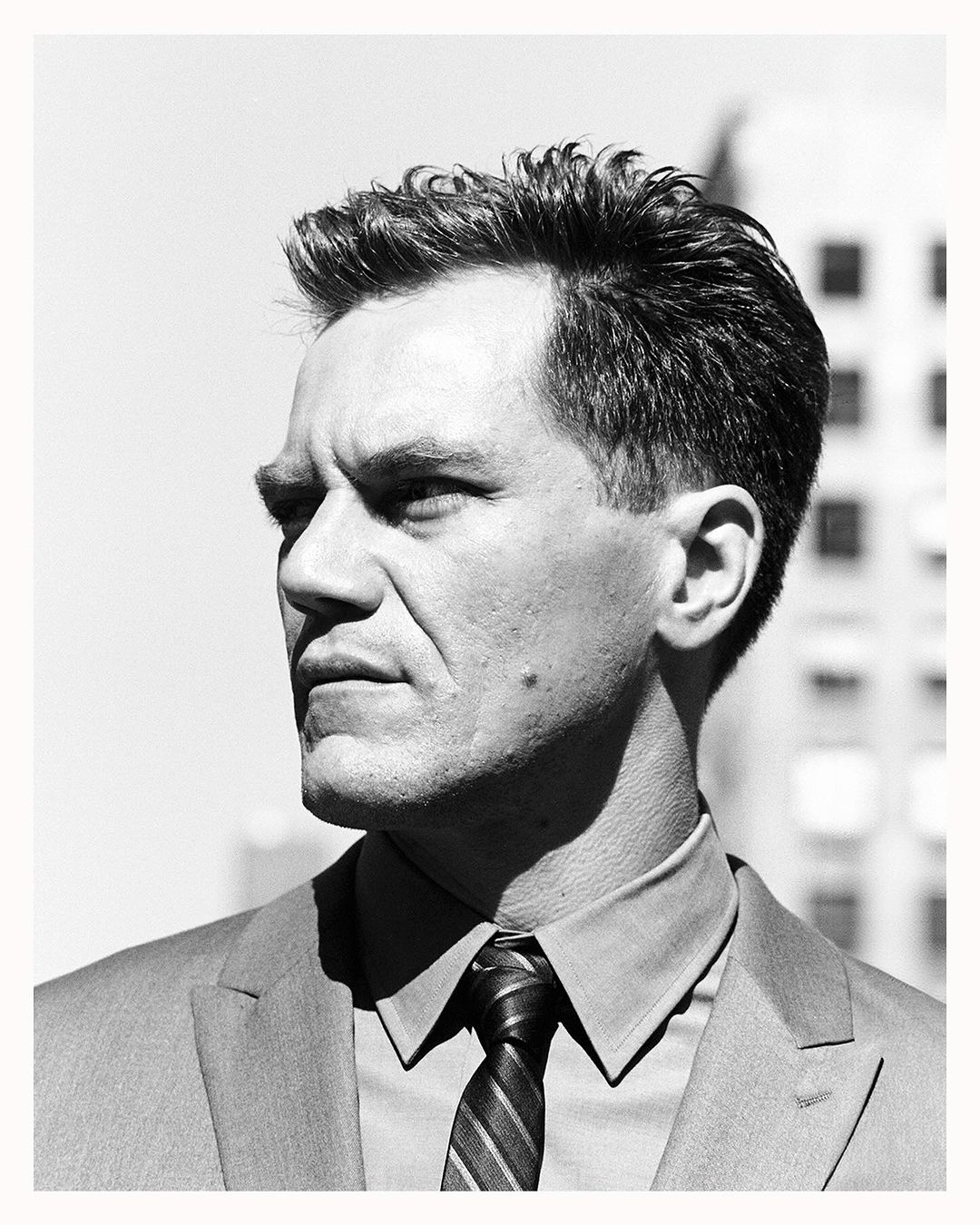 Monochrome portrait of an adult male wearing a suit and tie, standing outdoors with a tall building in the background on a bright day.