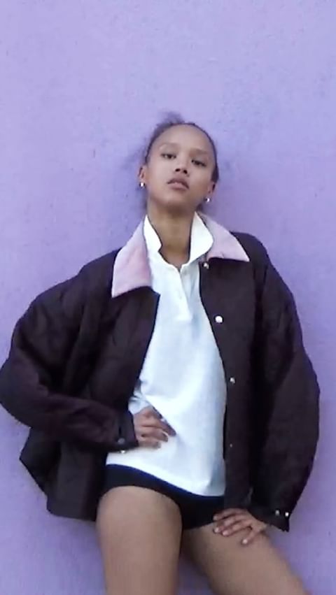 A young woman poses in a stylish brown jacket and white polo shirt against a solid light purple background in a studio setting.
