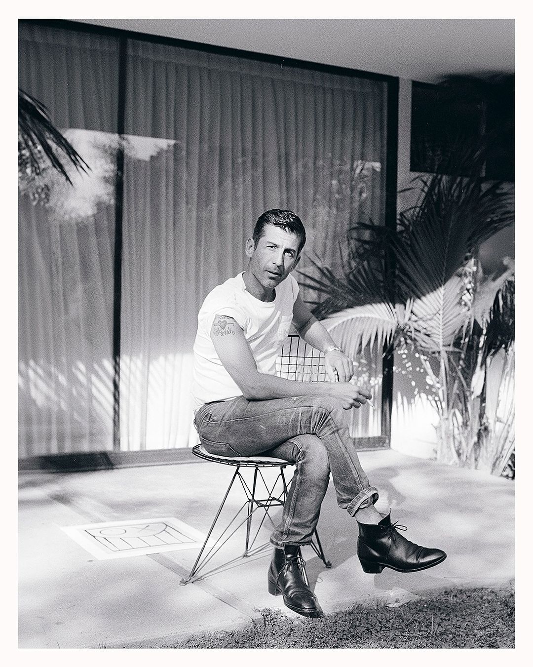 Black and white portrait of a man in a white t-shirt and jeans sitting outside on a wire chair, with a building and tropical plants.