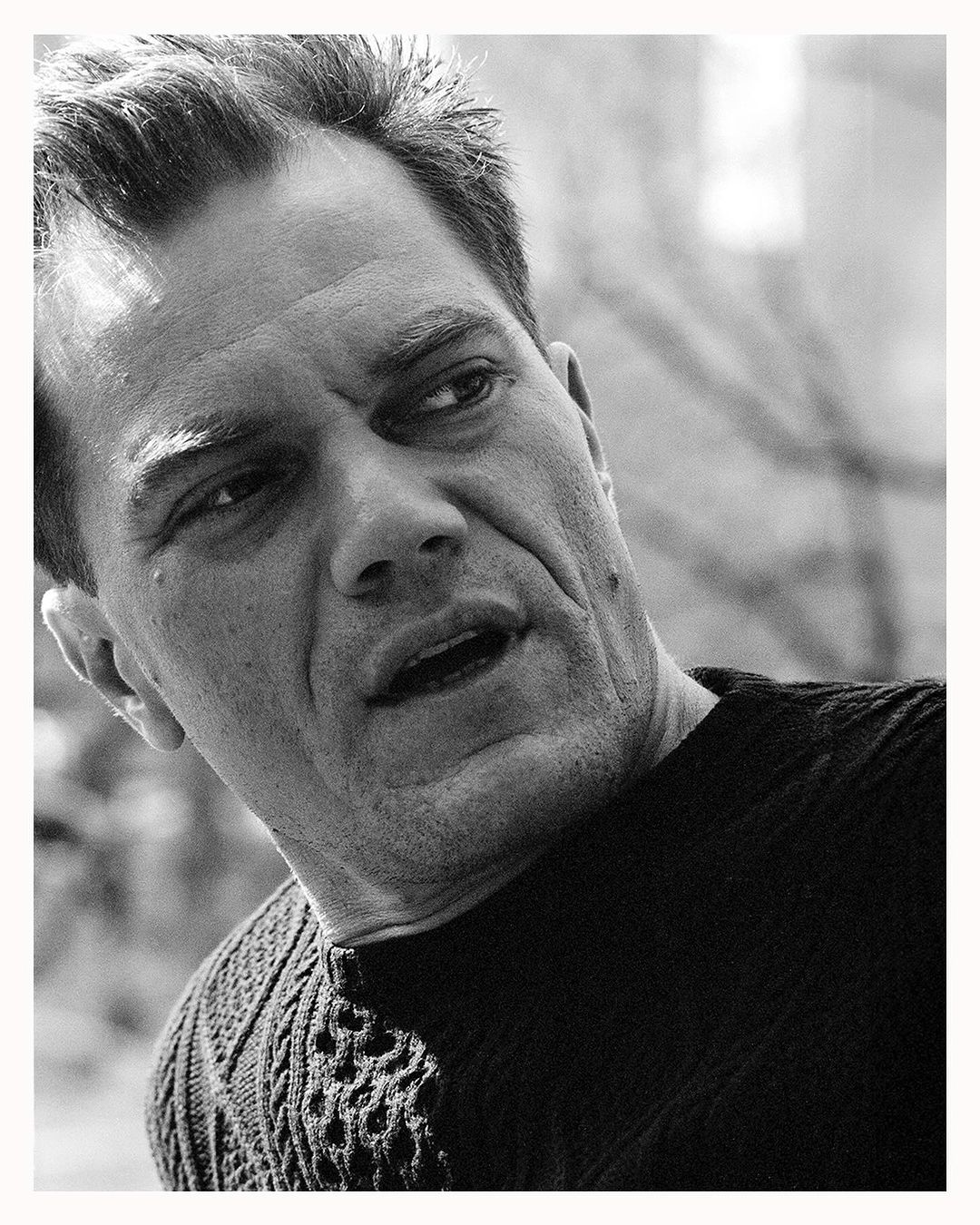 Black and white portrait of a man with short hair, wearing a dark patterned sweater, looking off to the side outdoors with natural light.