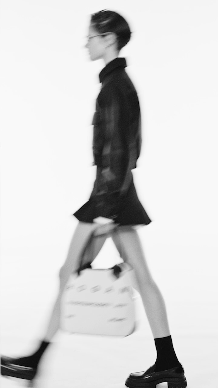 Woman in black jacket, skirt, socks, and platform shoes walking with a white handbag against a blurred white background.