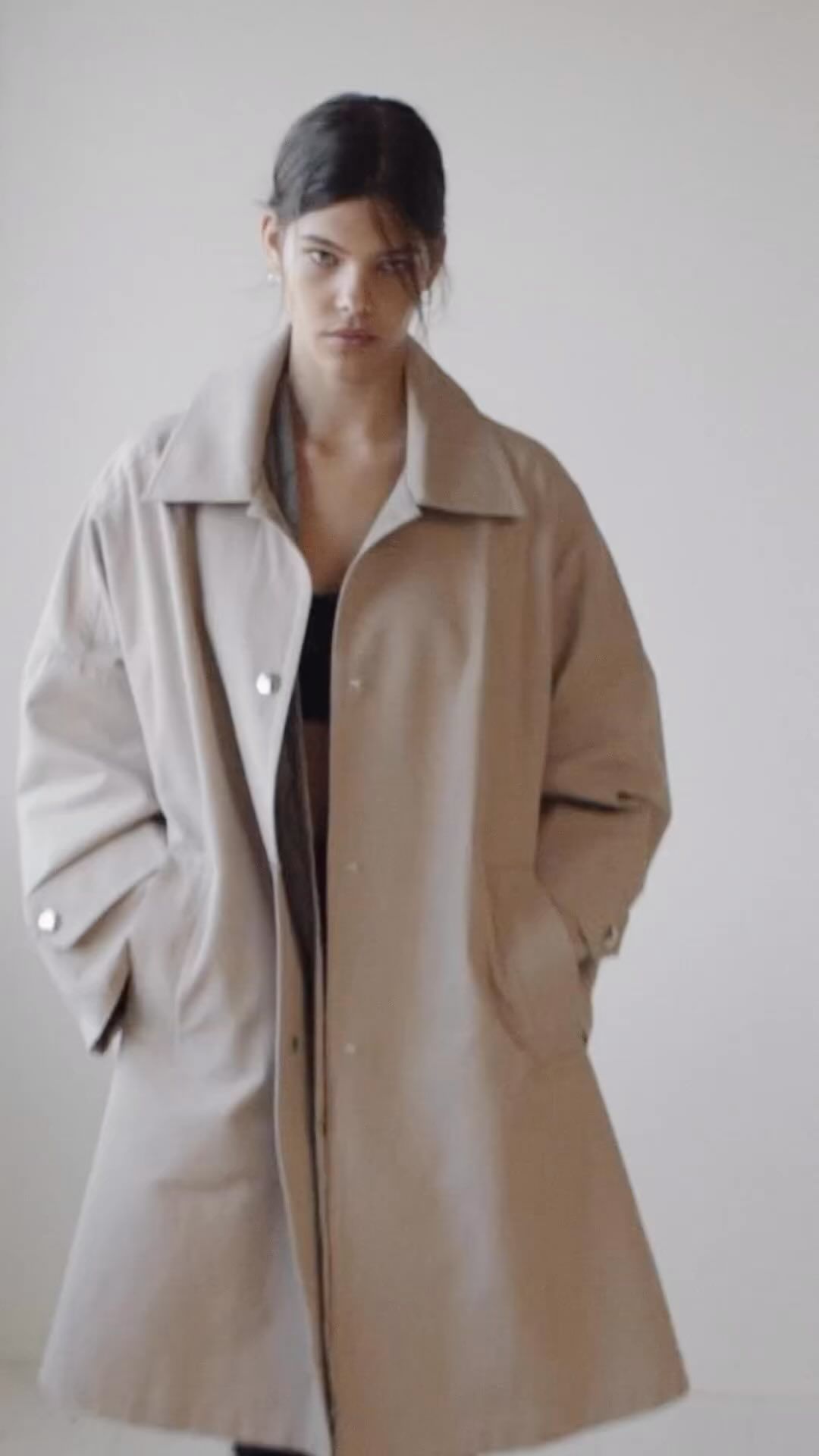 A fashionable adult woman is posing indoors wearing a light brown trench coat, with her hands casually placed in her pockets, in a studio setting.