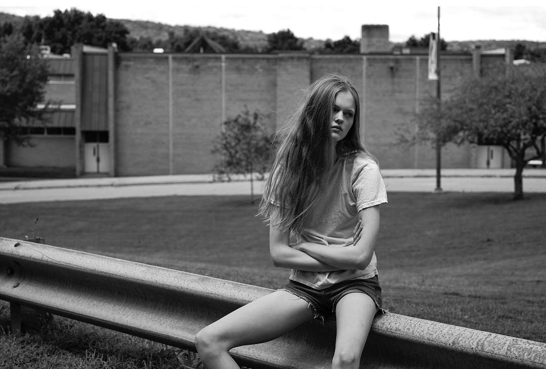 Black and white portrait of a young woman sitting on a guard rail with her arms crossed outdoors with a building and trees in the background.