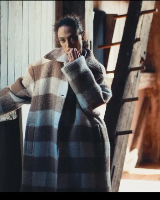 Adult female poses in a large tan and white plaid coat indoors, with a wooden wall and ladder in the background.