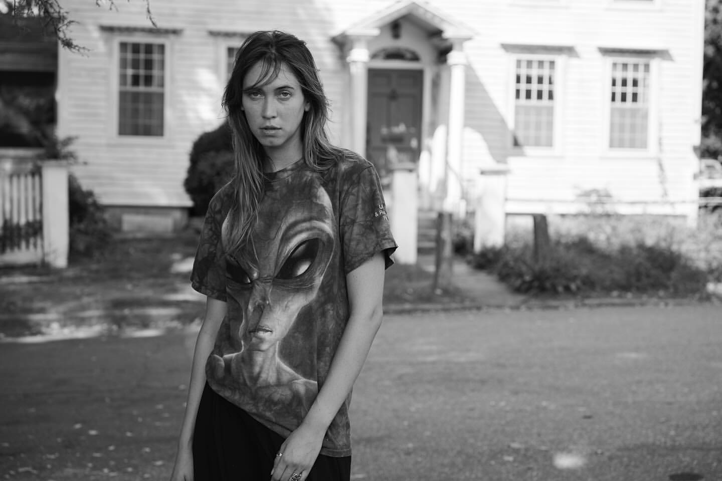 Monochrome portrait of a young adult female wearing an alien graphic tee shirt, standing in front of a white clapboard house outdoors.