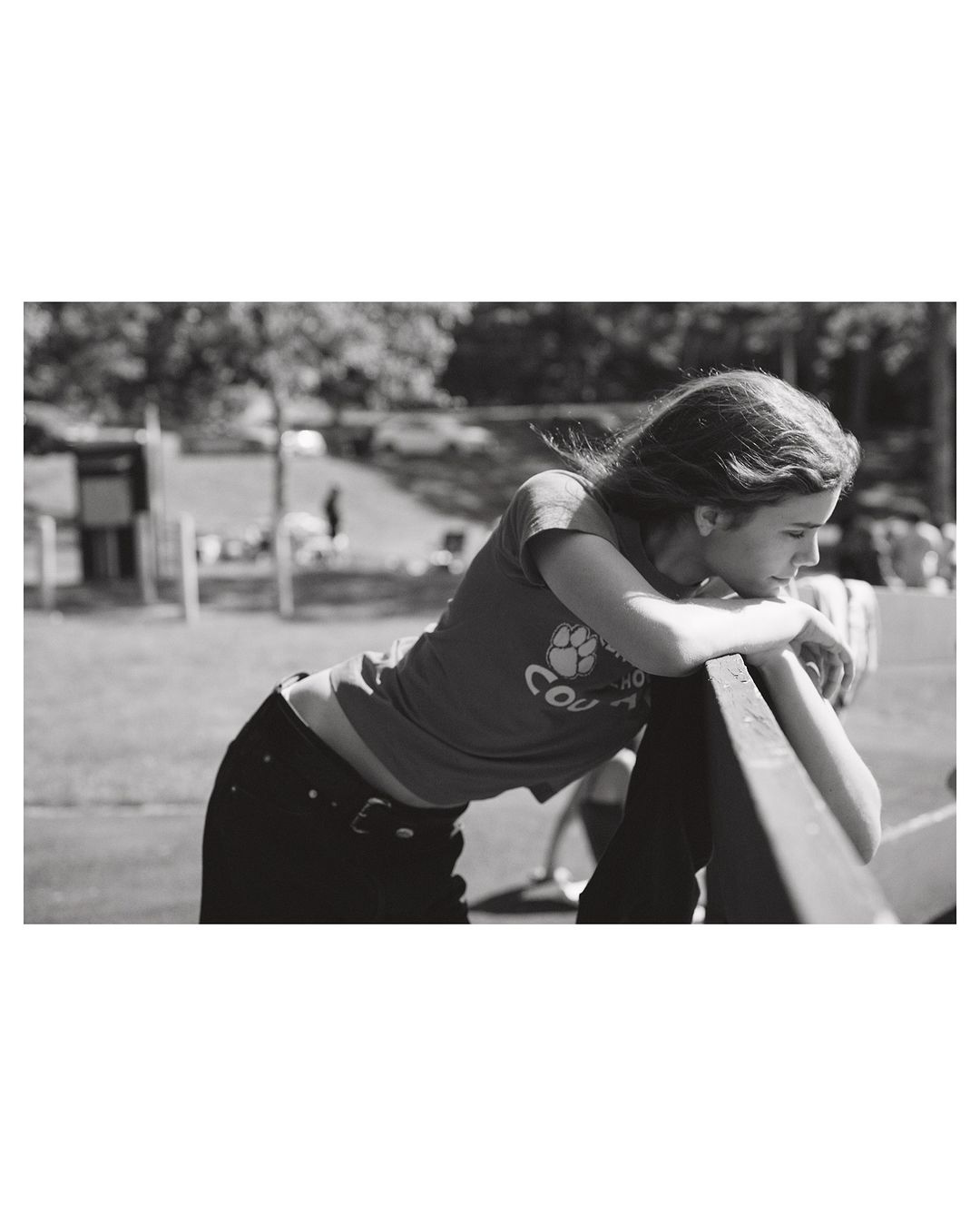 A young woman leans on a fence with a grassy field and trees in the background, shot in black and white with natural lighting.