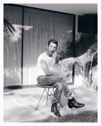 Monochrome portrait of a man in a white t-shirt and jeans seated on a wire chair outside a building.