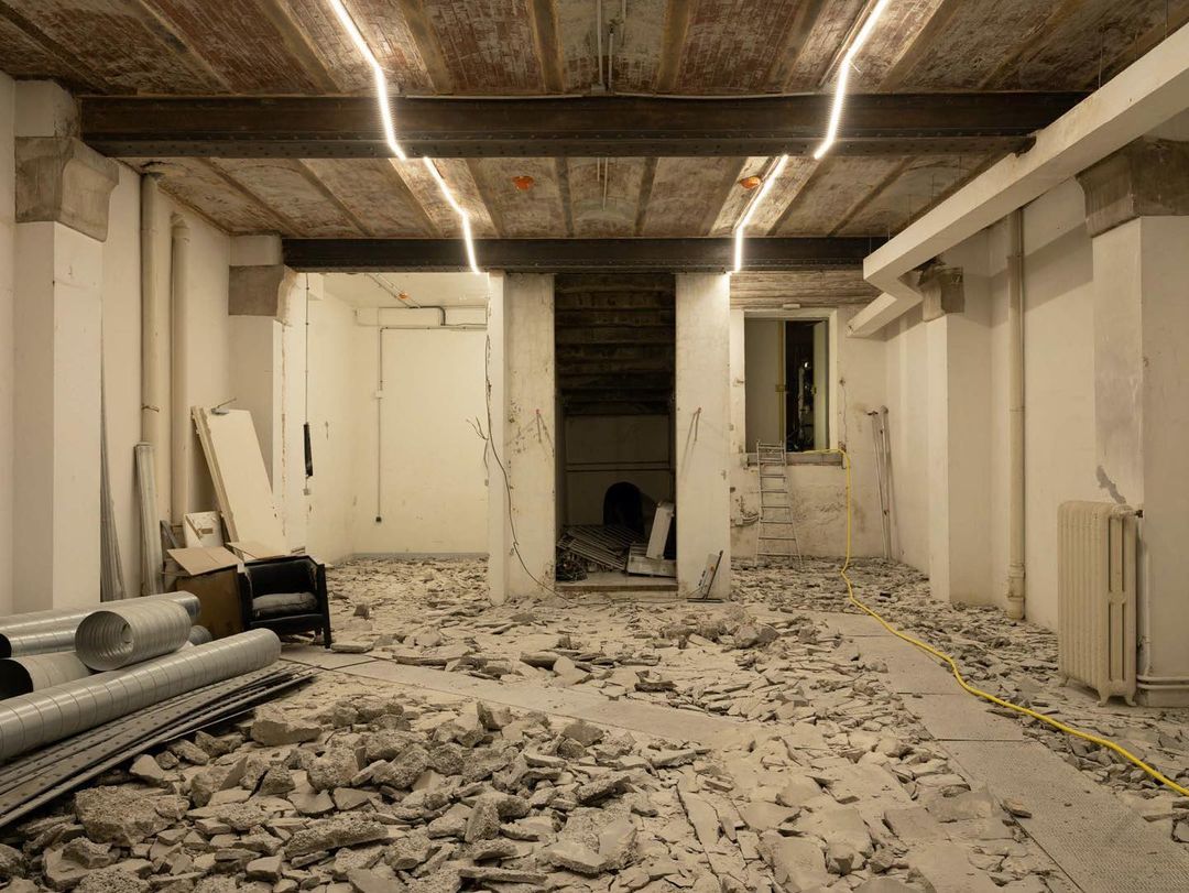Interior view of a construction site featuring debris-covered floors, exposed ceiling beams, and an open doorway, suggesting renovation.