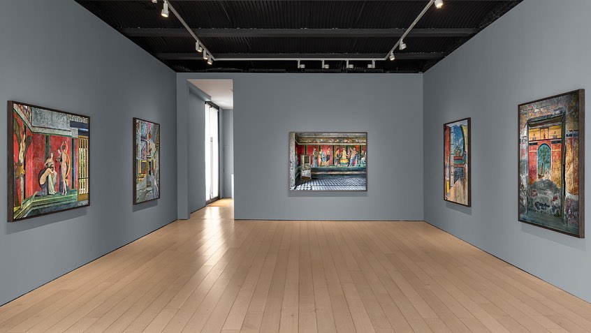 Art gallery featuring paintings with figural and architectural subjects hanging on gray walls, illuminated by track lighting.