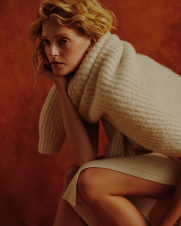A fair-skinned woman with curly blonde hair poses indoors wearing a chunky knit sweater and neutral skirt against a terracotta background.