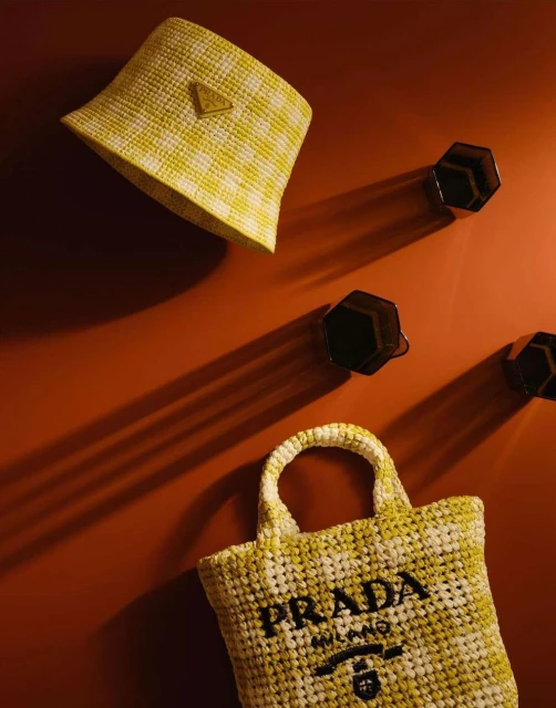 Still life of a yellow and white checkered Prada bucket hat and tote bag against a red-orange wall with geometric wall hooks.