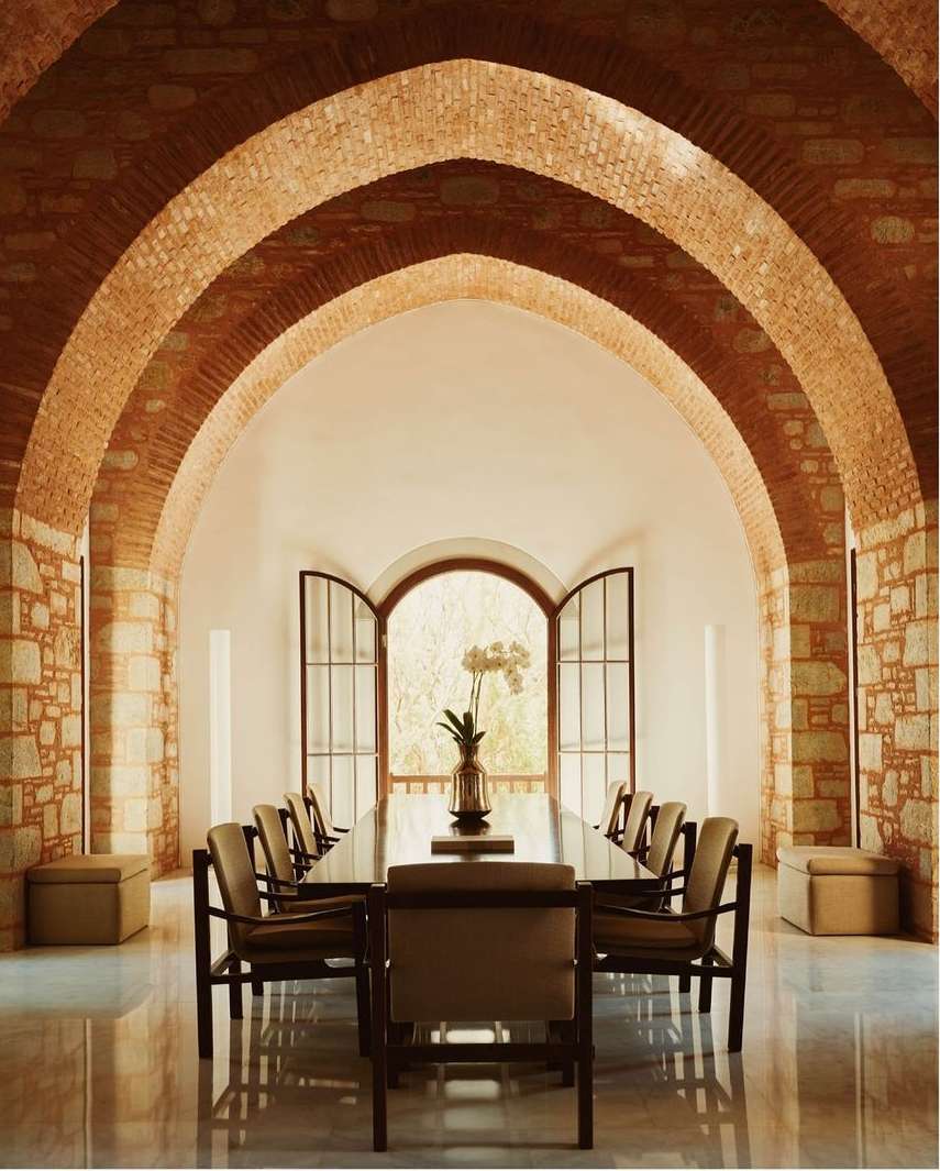 Indoor dining area featuring a long table with chairs, brick arches, and an open window, creating a bright and inviting space.